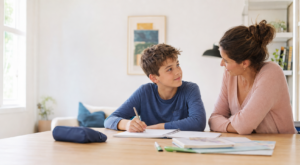 Un adolescent fait ses devoirs à table pendant qu'un parent l'observe avec attention.